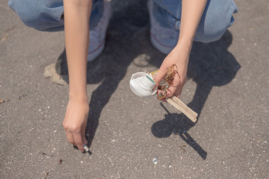 A person with light skin, dressed in light blue jeans and white sneakers, is crouching on a paved surface outdoors. They are holding a small, partially broken white ceramic object with a green stripe,